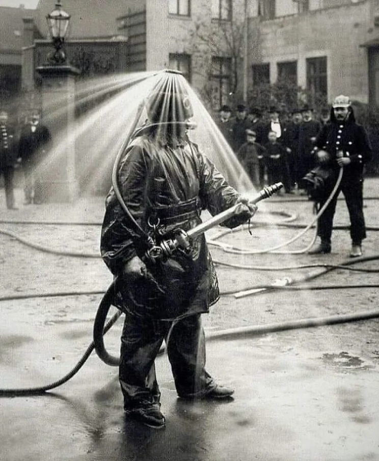 Historická fotografia hasiča v špeciálnom ochrannom obleku s vodným postrekom.