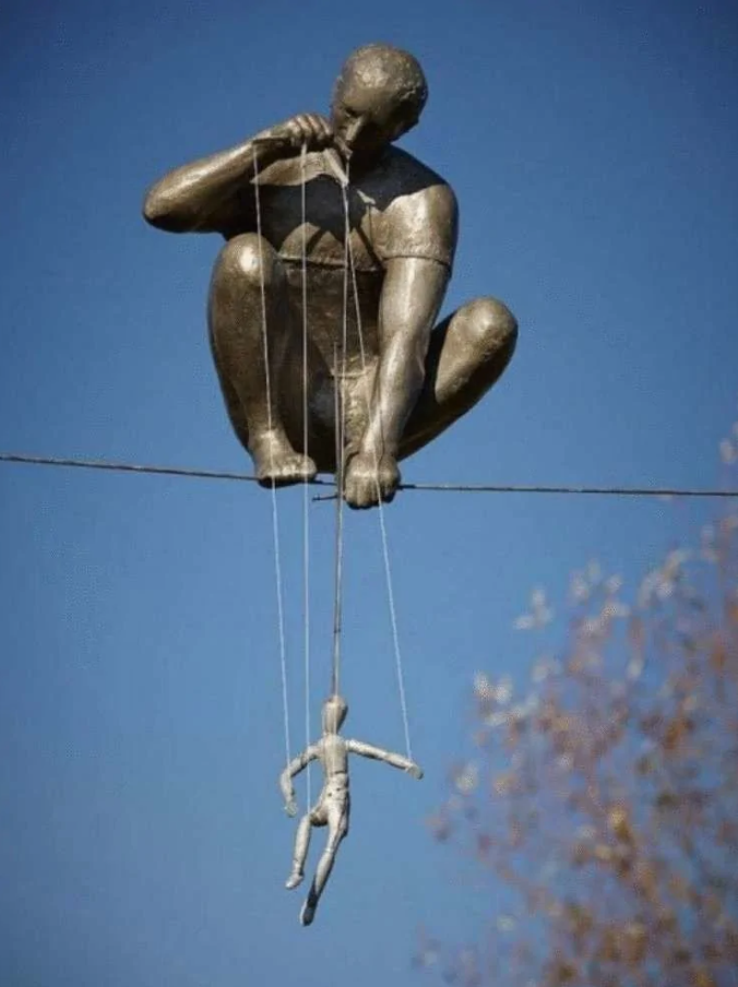 Socha muža ovládajúceho bábku na lane, symbolizujúca kontrolu a rovnováhu, s modrým nebom v pozadí.