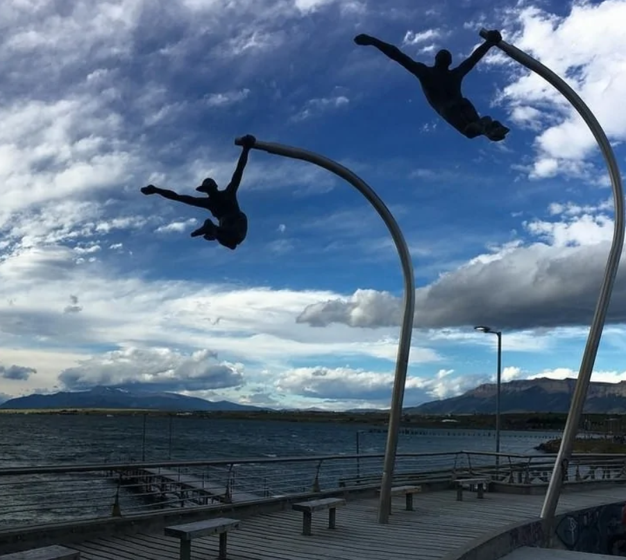 Sochy akrobatov na stĺpoch pri pobreží, proti dramatickej oblohe s horami v pozadí.
