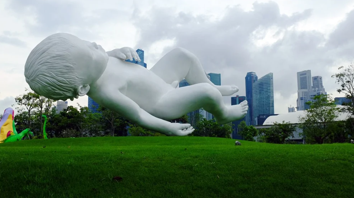 Obrovská socha dieťaťa leží na tráve v parku s mestským panorámovým pozadím.