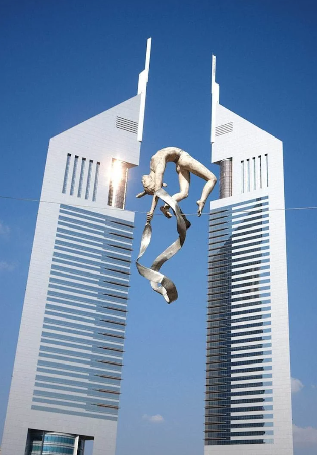Socha akrobata medzi modernými mrakodrapmi za jasného dňa, modrá obloha.