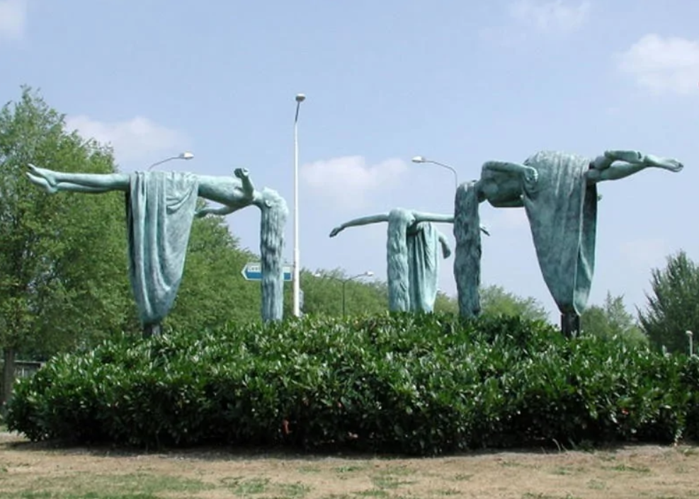 Bronzové sochy dráhových tanečníkov vo vzduchu, obklopené zeleňou na verejnom priestranstve.
