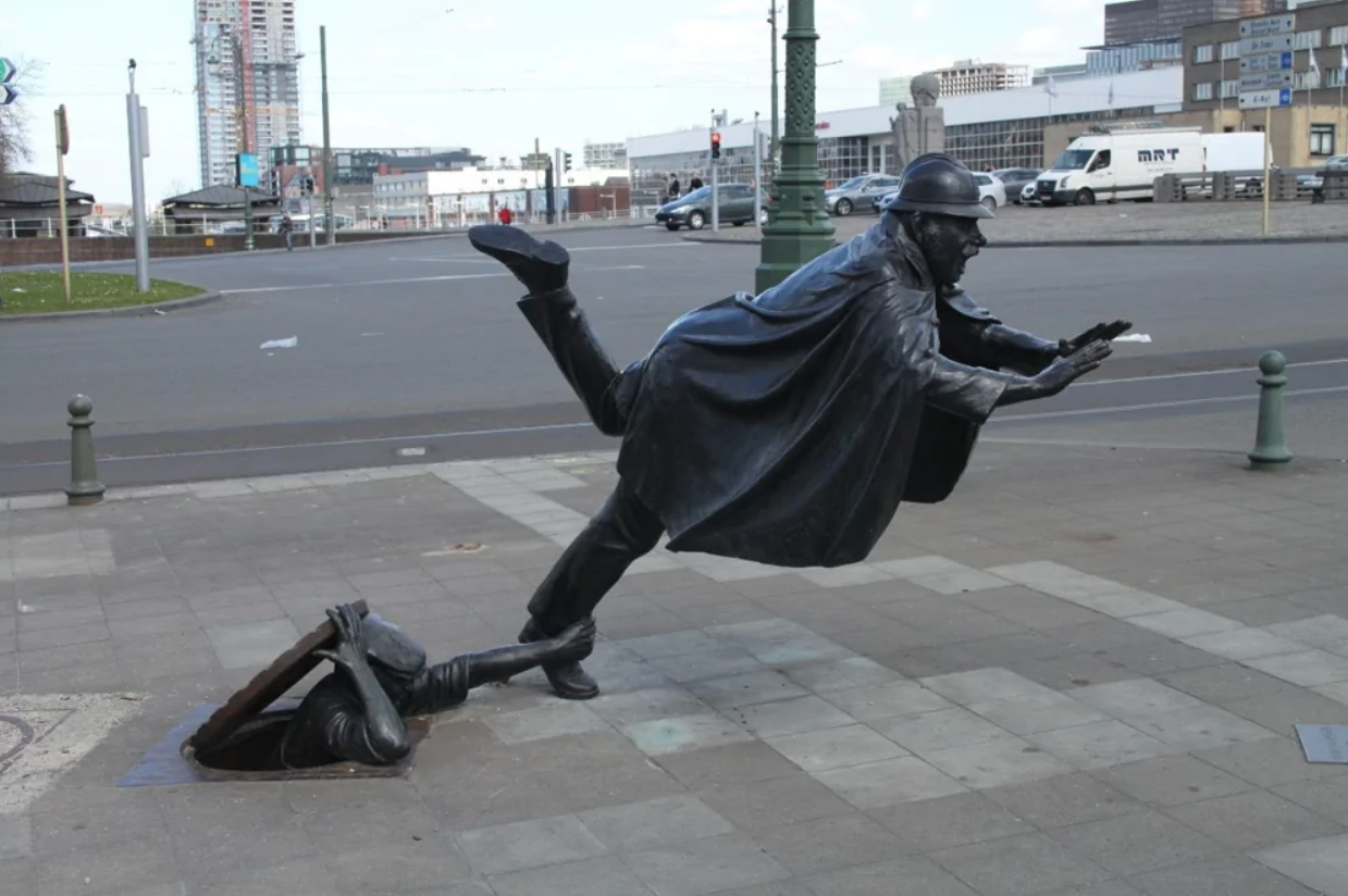 Bronzová socha človeka padá do kanála, ulicu zachytáva karikatúrna scéna.