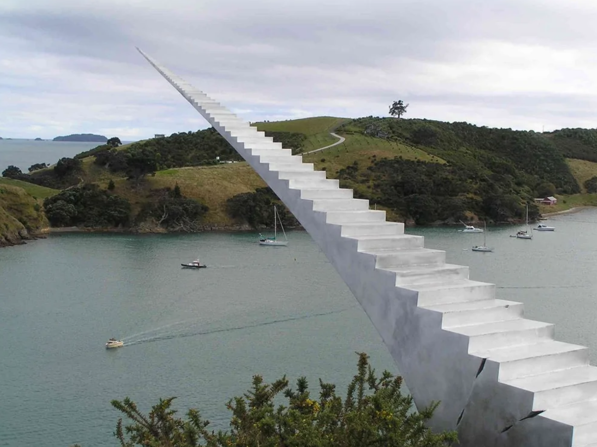 Schody smerujúce do neba nad zálivom, obklopené kopcami a loďami na vode. Optická ilúzia v prírode.