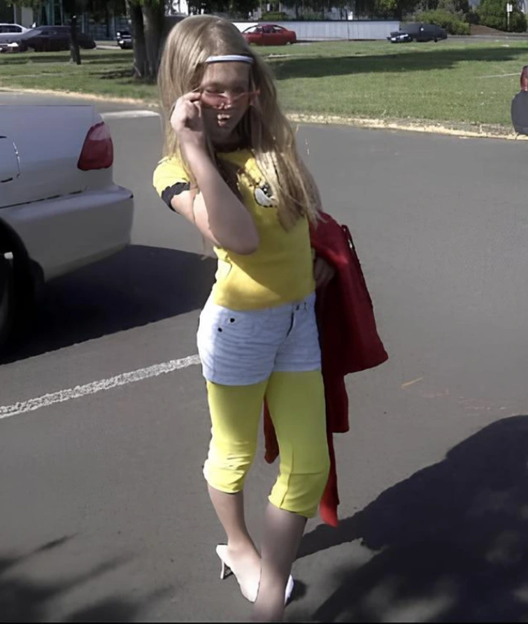 Dievča v žltom tričku a okuliaroch pózuje na parkovisku, drží červenú bundu a usmieva sa na kameru.