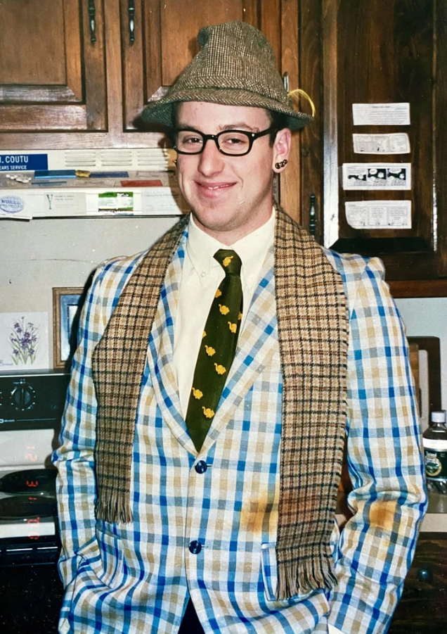 Muž v klobúku a károvanom saku s kravatou a šálom stojaci v kuchyni s drevenými skrinkami.