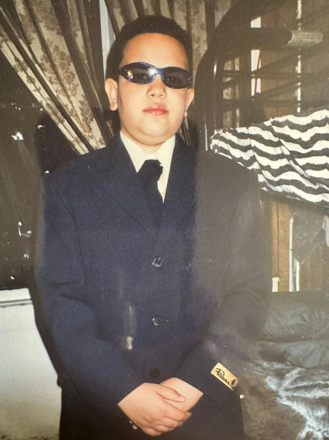 Chlapec v obleku a slnečných okuliaroch stojí pred závesom, s rukami spojenými pred sebou.