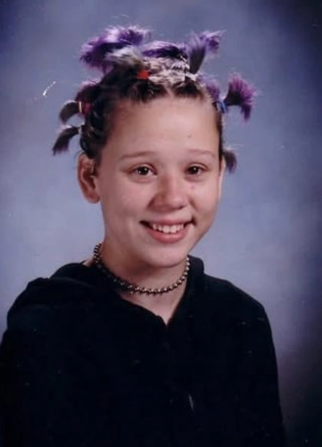 Mladá osoba s farebnými vrkočmi na tmavom pozadí, oblečená v čiernom a s úsmevom na tvári.