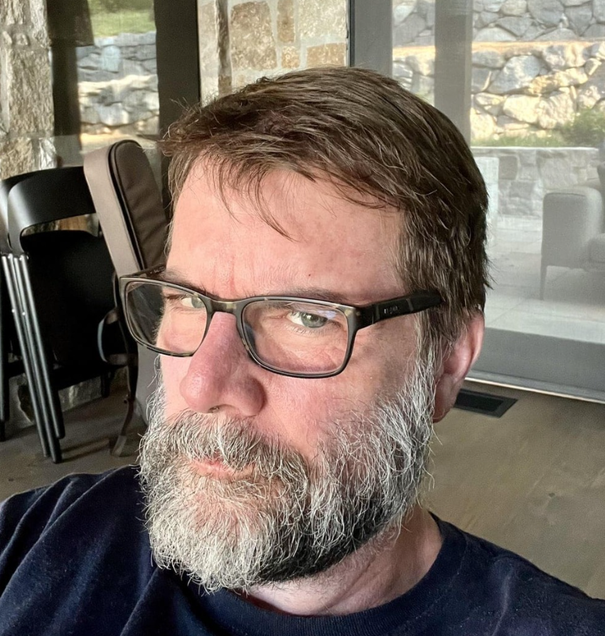 osoba so sivou bradou a okuliarmi sedí v miestnosti so sklennými dverami a kamennou stenou