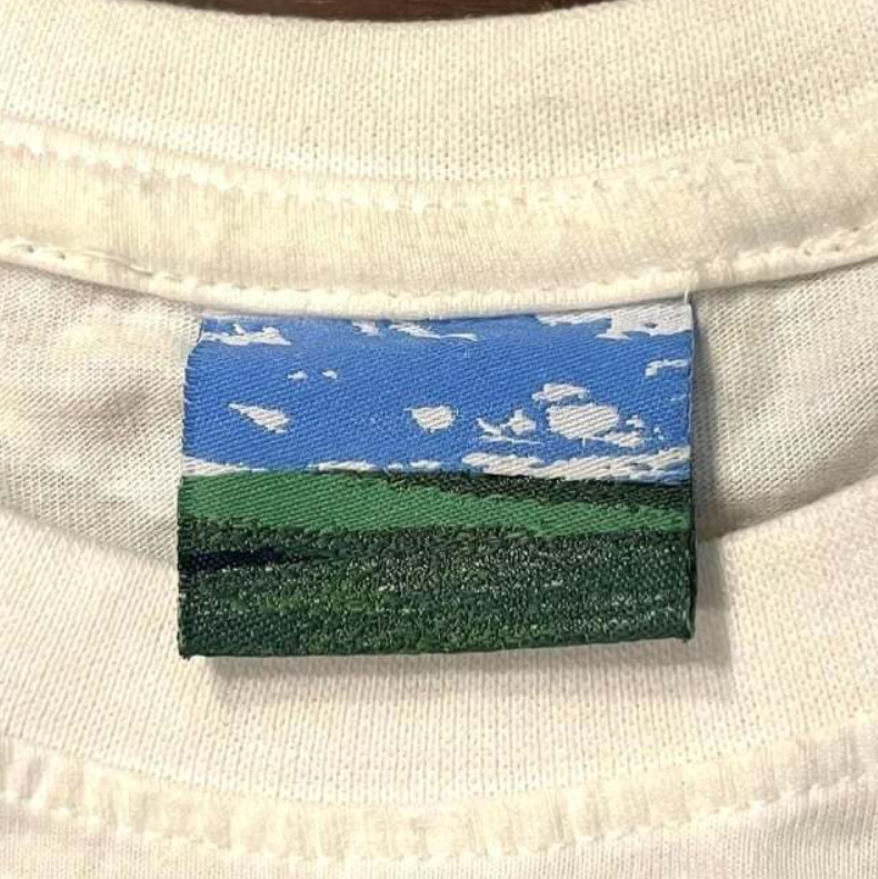 Biely tričko s visačkou, krajina zeleného poľa a modrej oblohy s oblakmi.