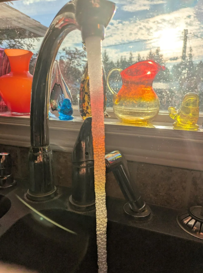 Tečúca voda z kohútika s farebnými nádobami na okne, slnečné svetlo preniká do kuchyne.