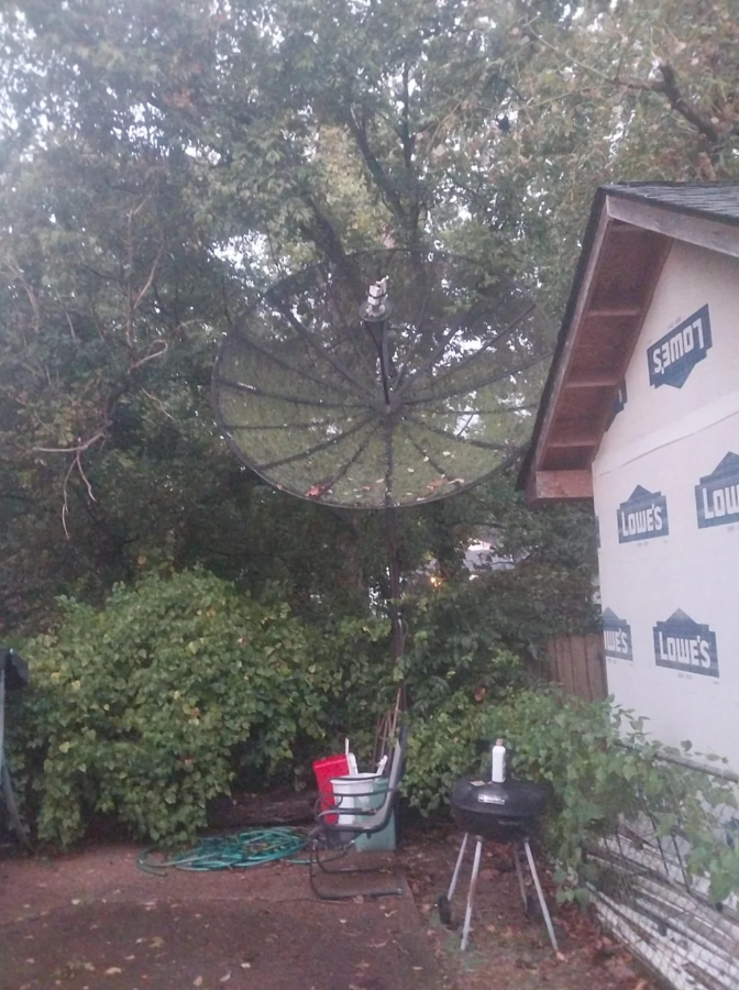 Stará satelitná anténa pri dome s altánkom, obklopená stromami a záhradným náčiním.