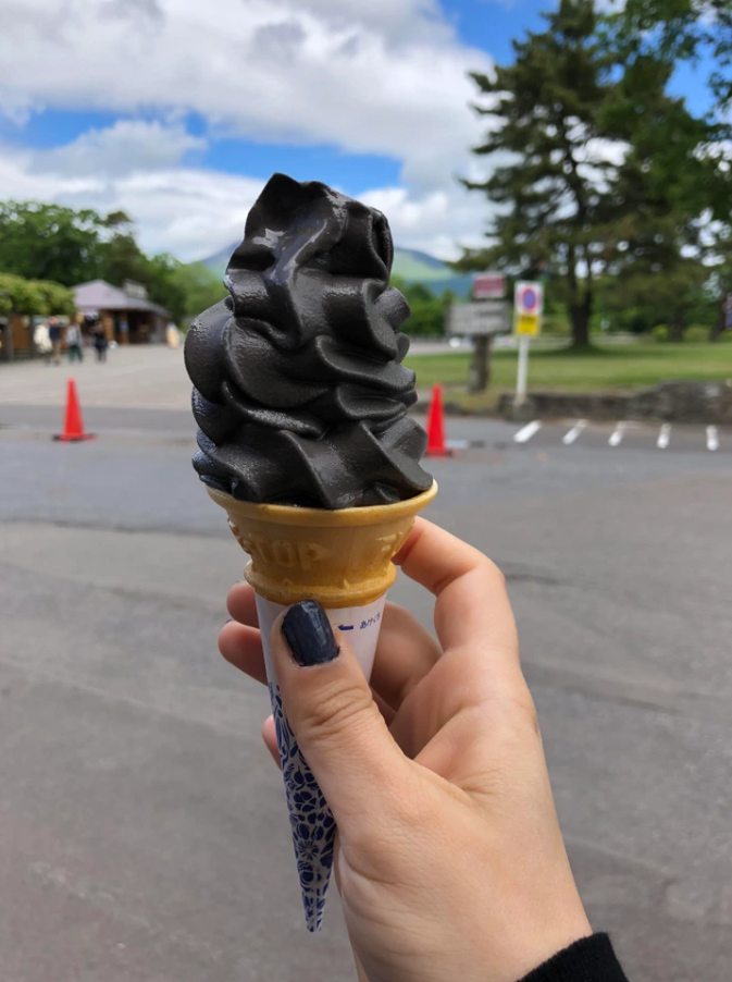 Osoba drží kužeľ zmrzliny s čiernou príchuťou na vonkajšom pozadí parkoviska a stromov.