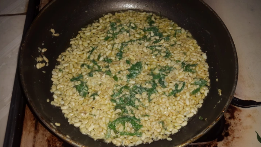 Miešaná zmes v panvici, obsahuje zelené listy a žlté zrná, varenie na sporáku.