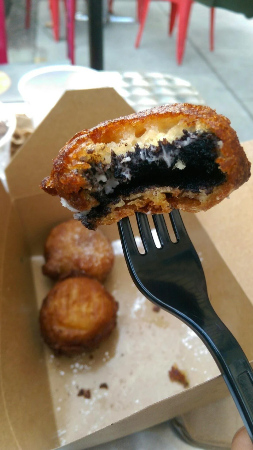 Vyprážané Oreo na plastovej vidličke, zlatisté cesto odhaľuje tmavú sušienku s krémovou náplňou.