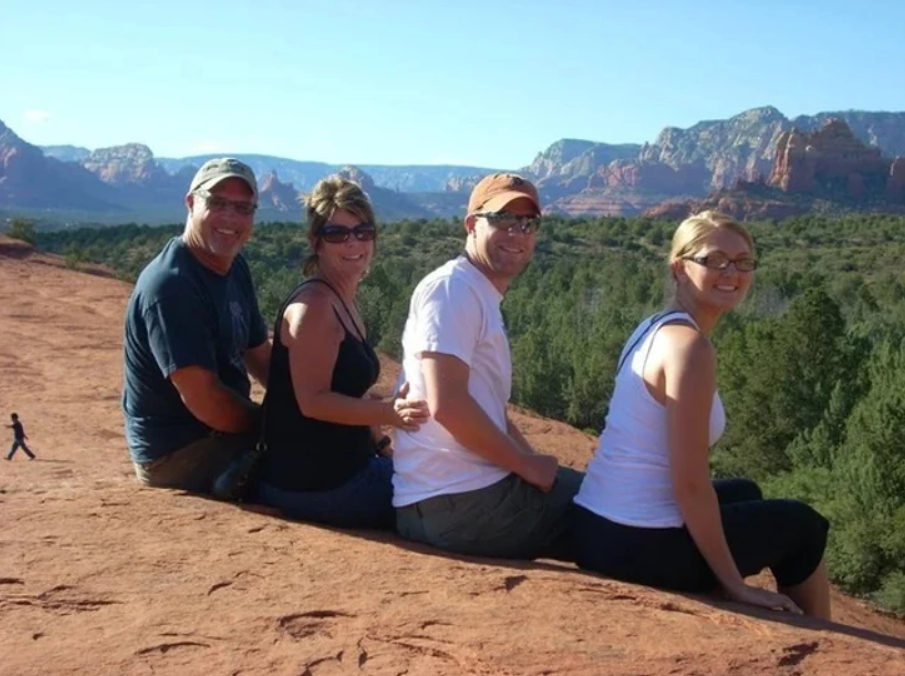 Bohužiaľ, neviem, kto sú ľudia na fotografii. Vidím však, že posedávajú na skalnom útese s výhľadom na les a hory v pozadí.