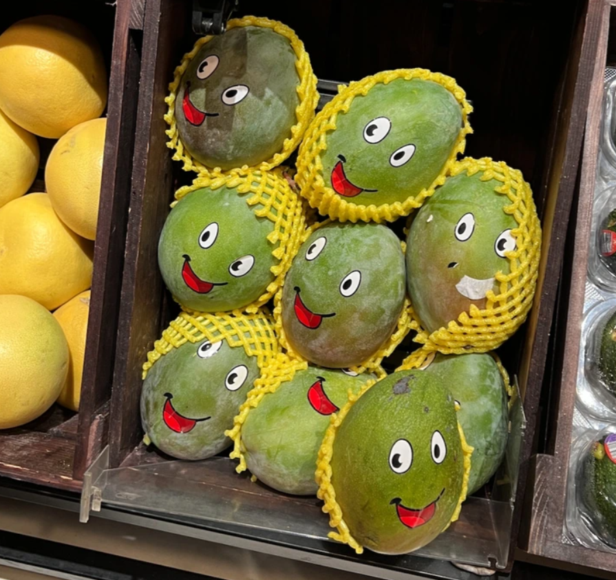 Mango s kreslenými tvárami v žltých sieťkach vedľa grapefruitov v obchode s potravinami.