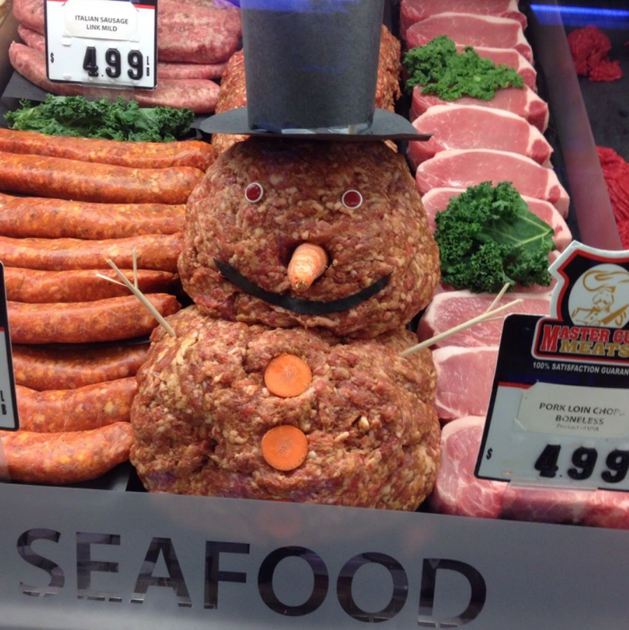 Snehuliak vytvorený z mäsovej zmesi, ozdobený klobúkom a mrkvou, v mäsovom oddelení s mäsovými výrobkami v pozadí.