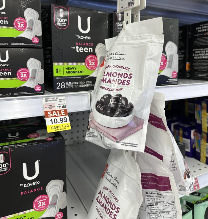 Balíky mandlí v tmavej čokoláde zavesené na poličke vedľa vložiek Kotex v obchode.
