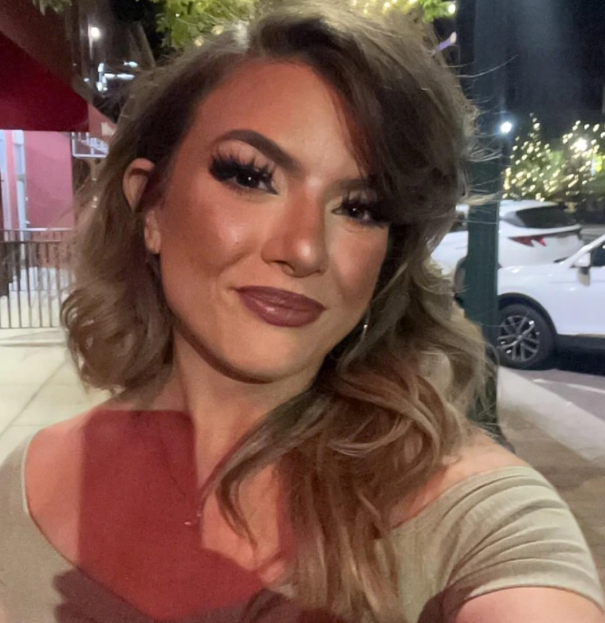 Osoba s dlhými vlasmi usmievajúca sa na nočnej ulici, osvetlená pouličnými lampami, vedľa zaparkovaného auta.