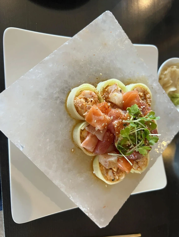 Luxusné sushi na štvorcovom tanieri ozdobené zelenými bylinami a ružovým lososom, servírované na ľadovej platni.