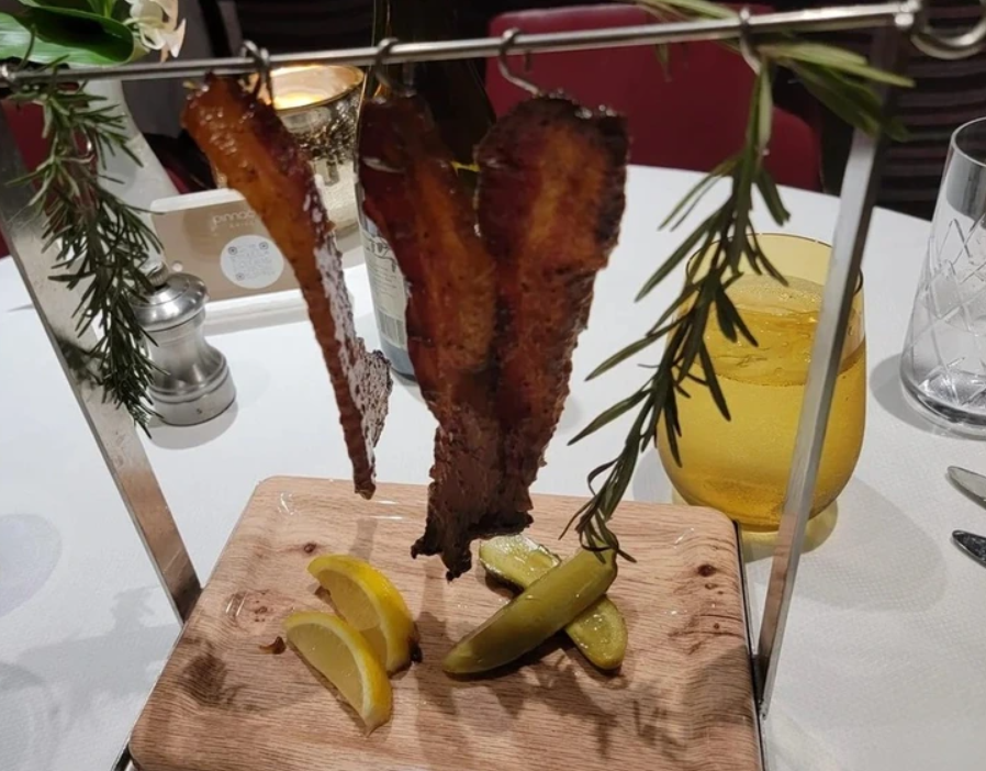 Kreatívne servírovanie slaniny na stojane s vetvičkami rozmarínu, citrónmi a uhorkami na drevenej doske.