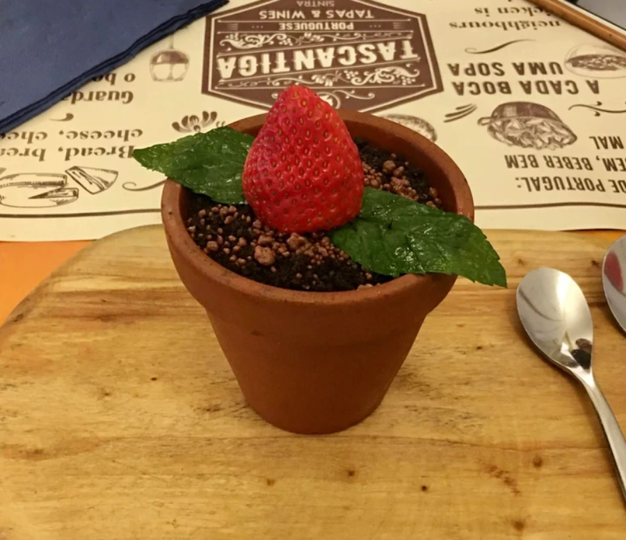 Dezert v kvetináči s čokoládovými kúskami, ozdobený jahodou a mätovým lístkom na drevenej doske.