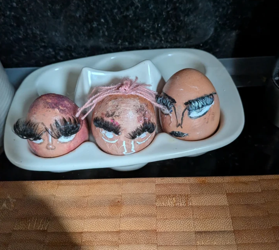 Tri vajíčka s maľovanými tvárami a mihalnicami v stojane na vajíčka na kuchynskej doske.