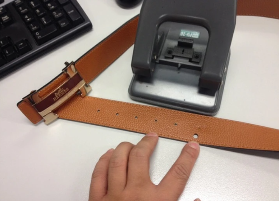 Opasok s novou dierkou pri dierovači na stole, vedľa klávesnice a ruky osoby.