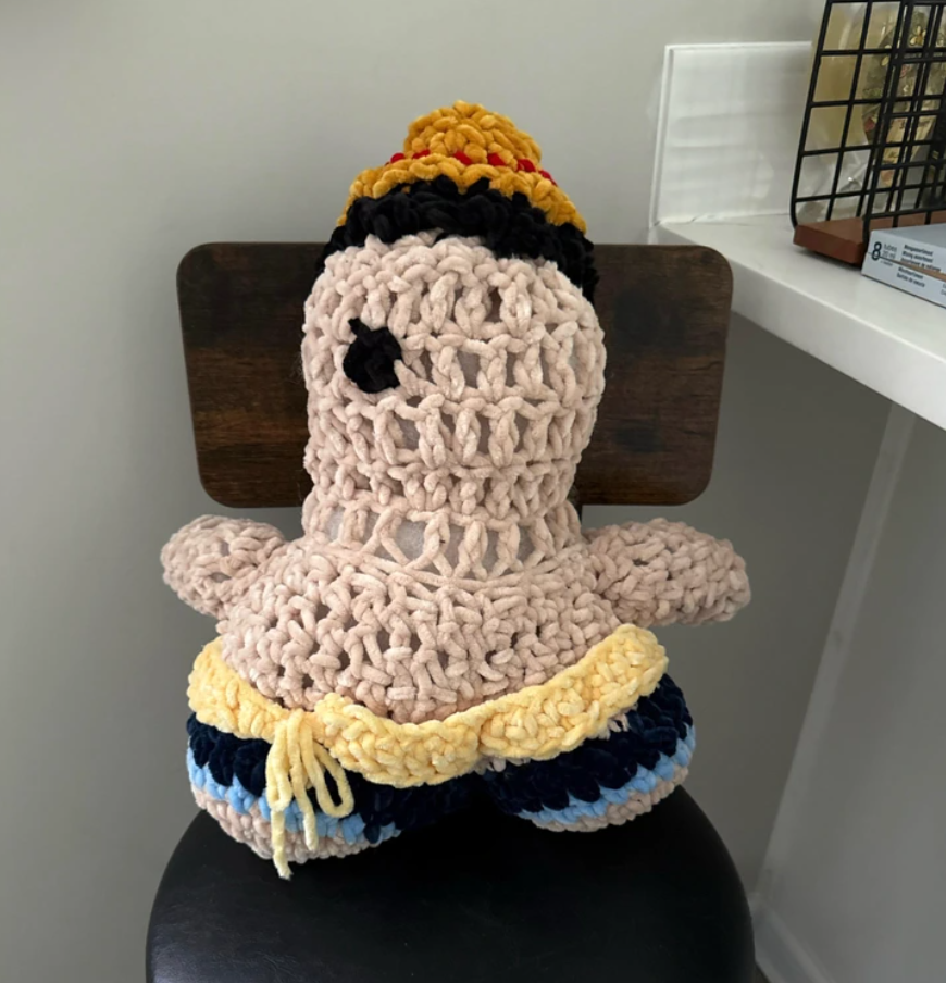 Háčkovaná bábika sediaca na stoličke, oblečená v modro-žltých šatách s čiernymi vlasmi a čiapkou.