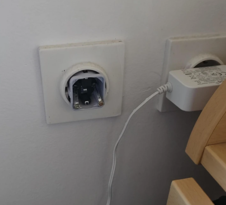 Zásuvka bez krytu s nabíjačkou na stene vedľa schodov, detailná fotografia elektrického pripojenia.