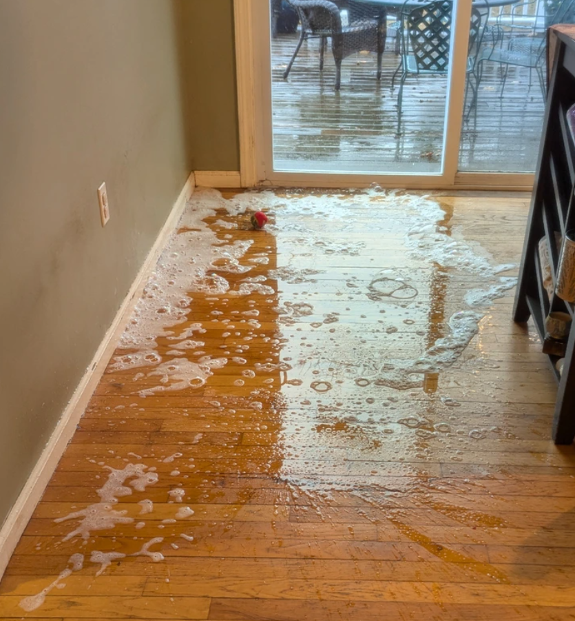 Voda s penou na podlahe pri sklenených dverách, pravdepodobne po daždi alebo prasknutej hadici, vedľa červenej hračky.