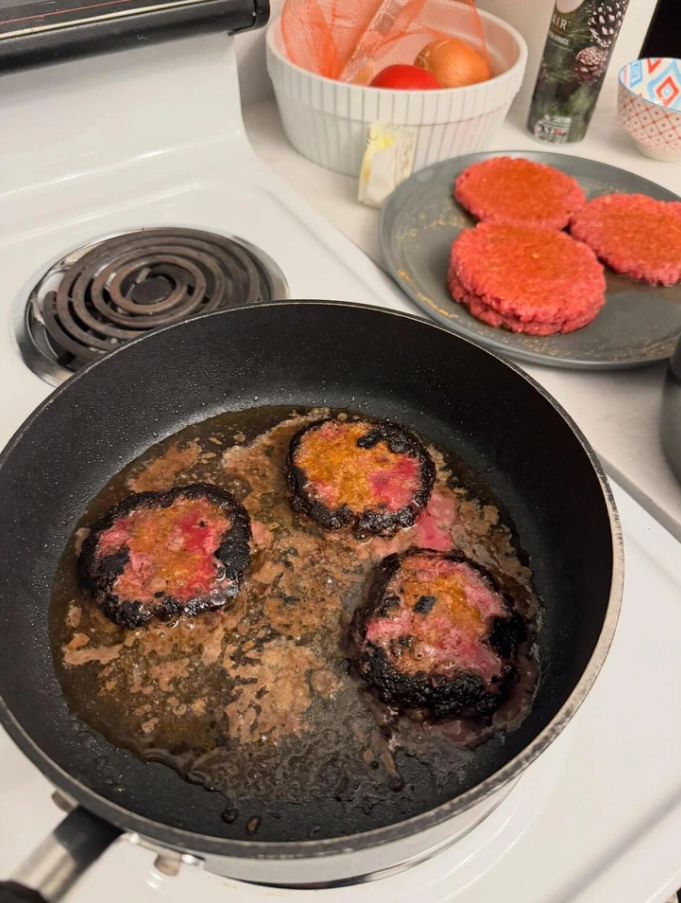 Hamburgery sa smažia na panvici, vedľa pripravené nespravované mäsové placky v kuchyni na bielom sporáku.