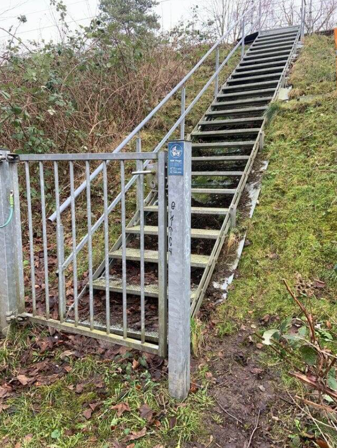 Kovový plot pred schodiskom vedúcim na mierny kopec s trávou a listami v pozadí.