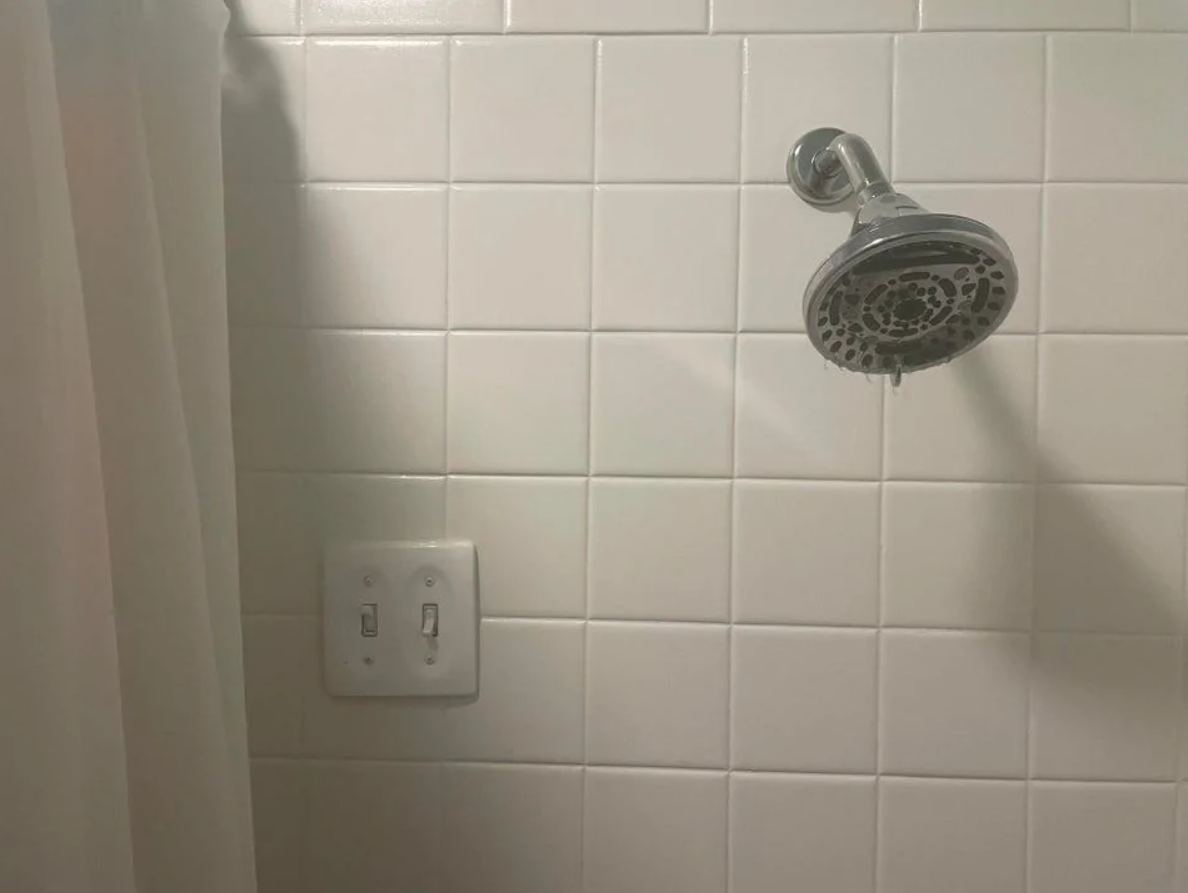 Sprchová hlavica na bielom obklade vedľa vypínačov, záves vedľa.