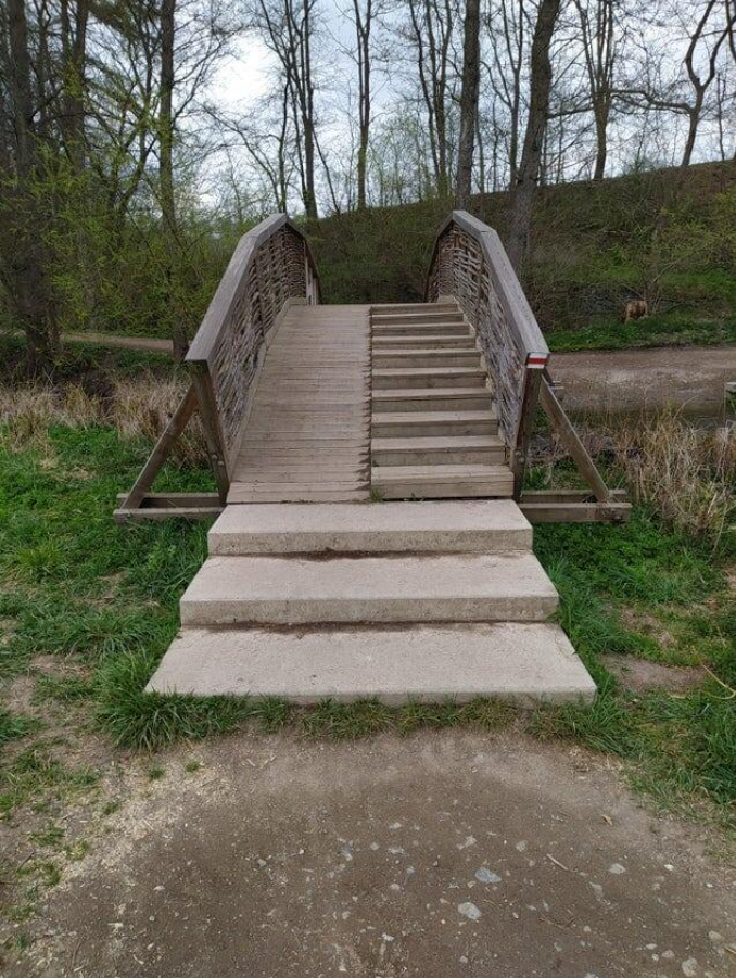 Drevený most cez potok s pevným zábradlím a schodmi, obklopený prírodou.