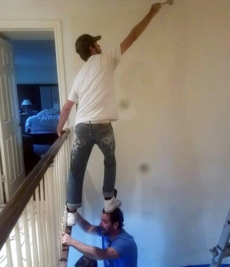 Dvaja muži maľujú stenu, pričom jeden stojí na druhom na schodišti.