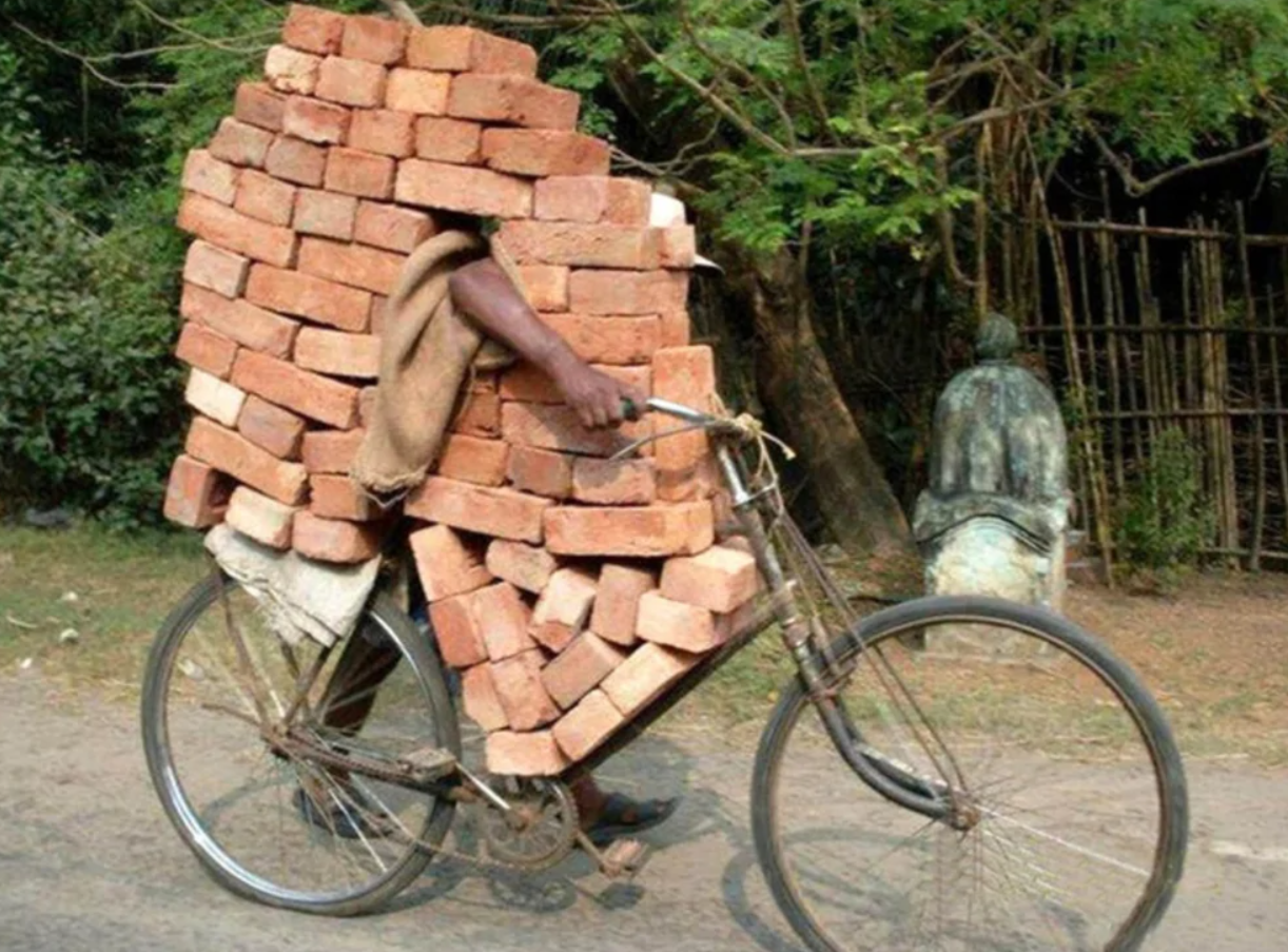 Muž nesúci množstvo tehál na bicykli po ceste, obklopený stromami.