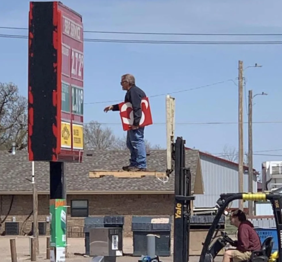 Muž nazdvihnutý vysokozdvižným vozíkom mení ceny na vonkajšej tabuli benzínky.