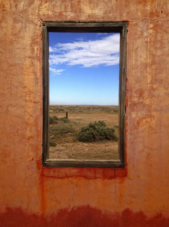 Staré okno v hlinenej stene ponúka výhľad na púštnu krajinu a modrú oblohu s oblakmi.