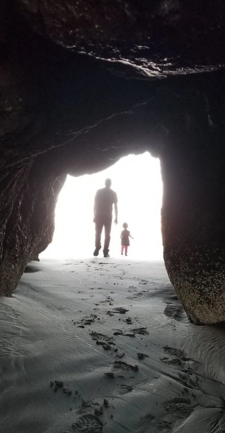 Dve siluetykráčajúce z jaskyne na pláž, jasné svetlo v pozadí zvyšuje tajomný pocit.