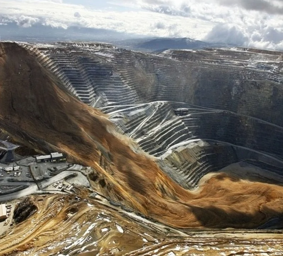 Veľká otvorená baňa s terasovitými svahmi a ťažobným zariadením na pozadí, obklopená horami a oblohou s oblakmi.