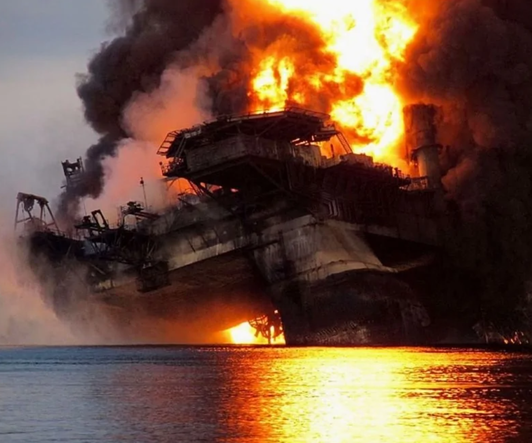 Horíaca ropná plošina na oceáne, šíriaci sa hustý dym a jasné plamene pri západe slnka nad vodnou hladinou.