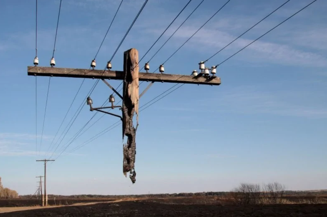 Poškodený elektrický stĺp s ohoreným kmeňom v prírodnej krajine pod modrou oblohou.