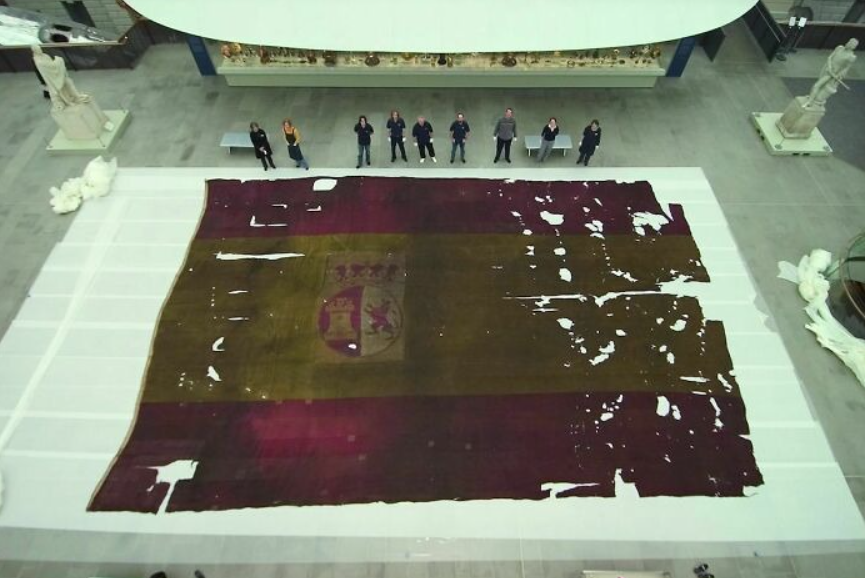 Veľká historická španielska vlajka s kormou leží na podlahe múzea, obklopená návštevníkmi a sochami.