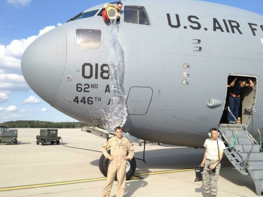 Letec pod kbelíkom vody vyliatym z prúdového lietadla na dráhe, ľudia sledujú scénu s úsmevom.