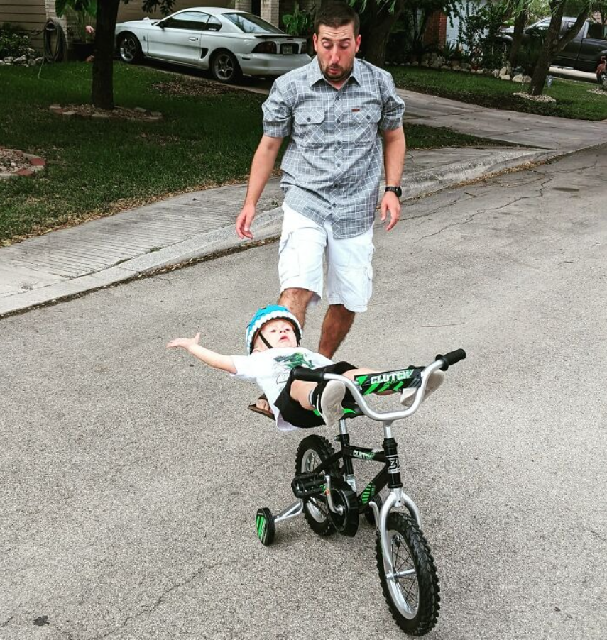Otec pomáha dieťaťu udržať rovnováhu na bicykli na ulici. Detská jazda sa nečakane skomplikovala.