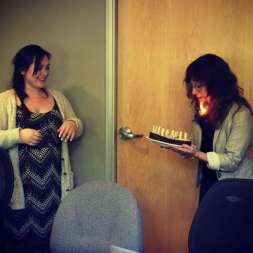 Žena drží tortu so sviečkami, zatiaľ čo druhá žena sa usmieva, v miestnosti s drevenými dverami.