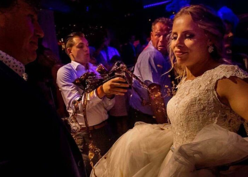 Usmiata nevesta v bielych šatách s rozliatym nápojom na svadobnej oslave s prekvapenými hosťami.