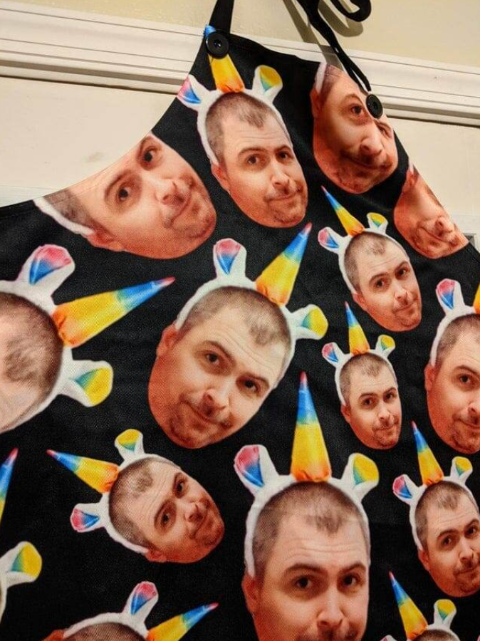 Vzorovaná zásterka s opakujúcimi sa tvárami a farebnými jednorožcami na čiernom pozadí.